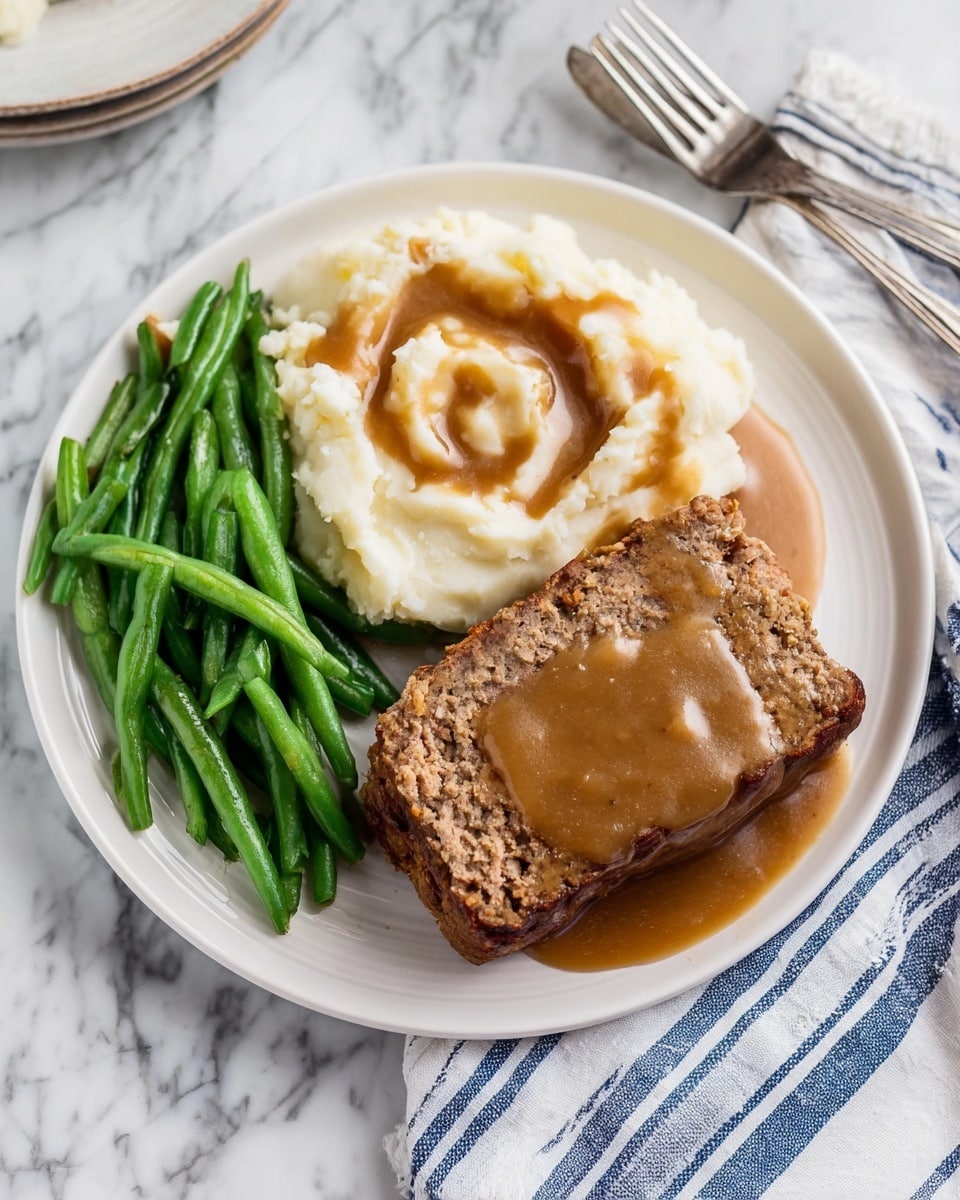 The dish on a white plate has three main parts: a pile of green beans on the left side, bright and slightly shiny with some butter melting on them; in the middle, a mound of creamy white mashed potatoes with a smooth texture, topped with a swirl of brown gravy; on the right side, a rectangular slice of meatloaf with a slightly rough texture, covered in the same brown gravy that pools a little on the plate. The plate is set on a white marbled surface with a fork resting on a blue and white striped napkin to the right. Photo taken with an iphone --ar 4:5 --v 7