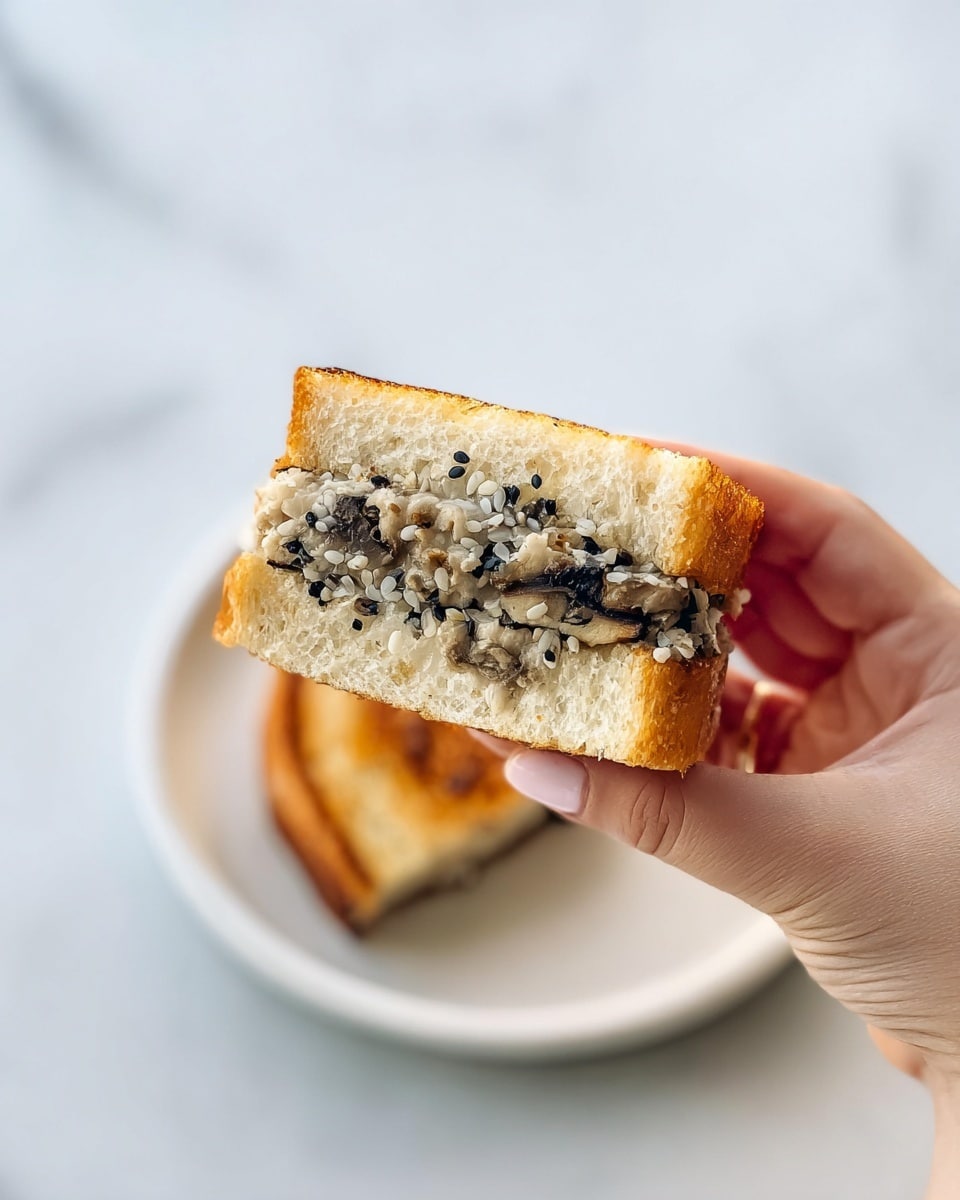 A close-up of a small sandwich held by a woman's hand showing three layers: the top and bottom layers are light golden-brown toasted bread with a slightly crispy texture, and the middle layer is a soft, moist filling composed of finely chopped black and beige ingredients, possibly mushrooms or vegetables, with small black sesame seeds scattered throughout. Behind the sandwich, on a white plate, is another piece of the sandwich with a similar toasted golden color. The scene is set on a white marbled surface, giving a clean and bright background. Photo taken with an iphone --ar 4:5 --v 7