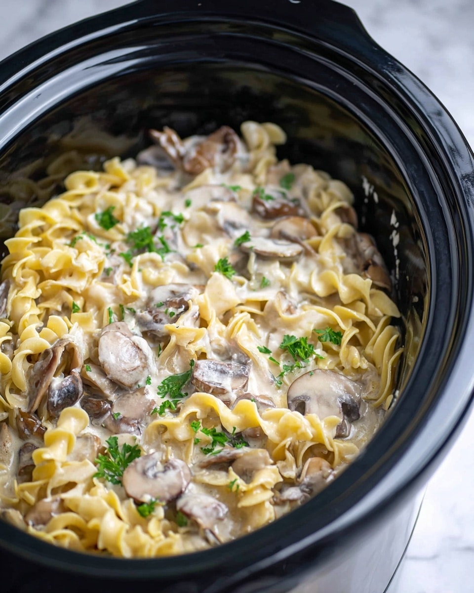 The image shows a close-up of a creamy noodle dish inside a shiny black slow cooker. The noodles are yellow and curly, layered with slices of light brown mushrooms and darker brown mushrooms, all coated in a thick, white creamy sauce. Small green parsley leaves are sprinkled on top, adding color contrast. The slow cooker has a reflective surface and is placed on a white marbled texture. Photo taken with an iphone --ar 4:5 --v 7