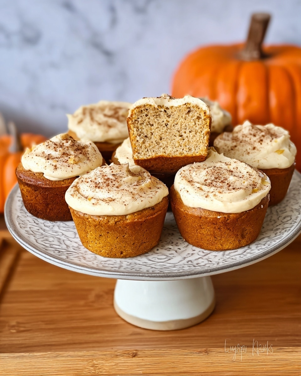 There are six small, round muffins on a decorative stand with a white base, each topped with a thick layer of creamy white frosting sprinkled with a fine dusting of cinnamon. One muffin is cut in half, showing a dense, moist, light beige interior. The stand is placed on a wooden surface, with a small orange pumpkin-shaped container in the background, all set against a white marbled texture. The muffins have a golden brown exterior with slight darker spots where baked. Photo taken with an iphone --ar 4:5 --v 7