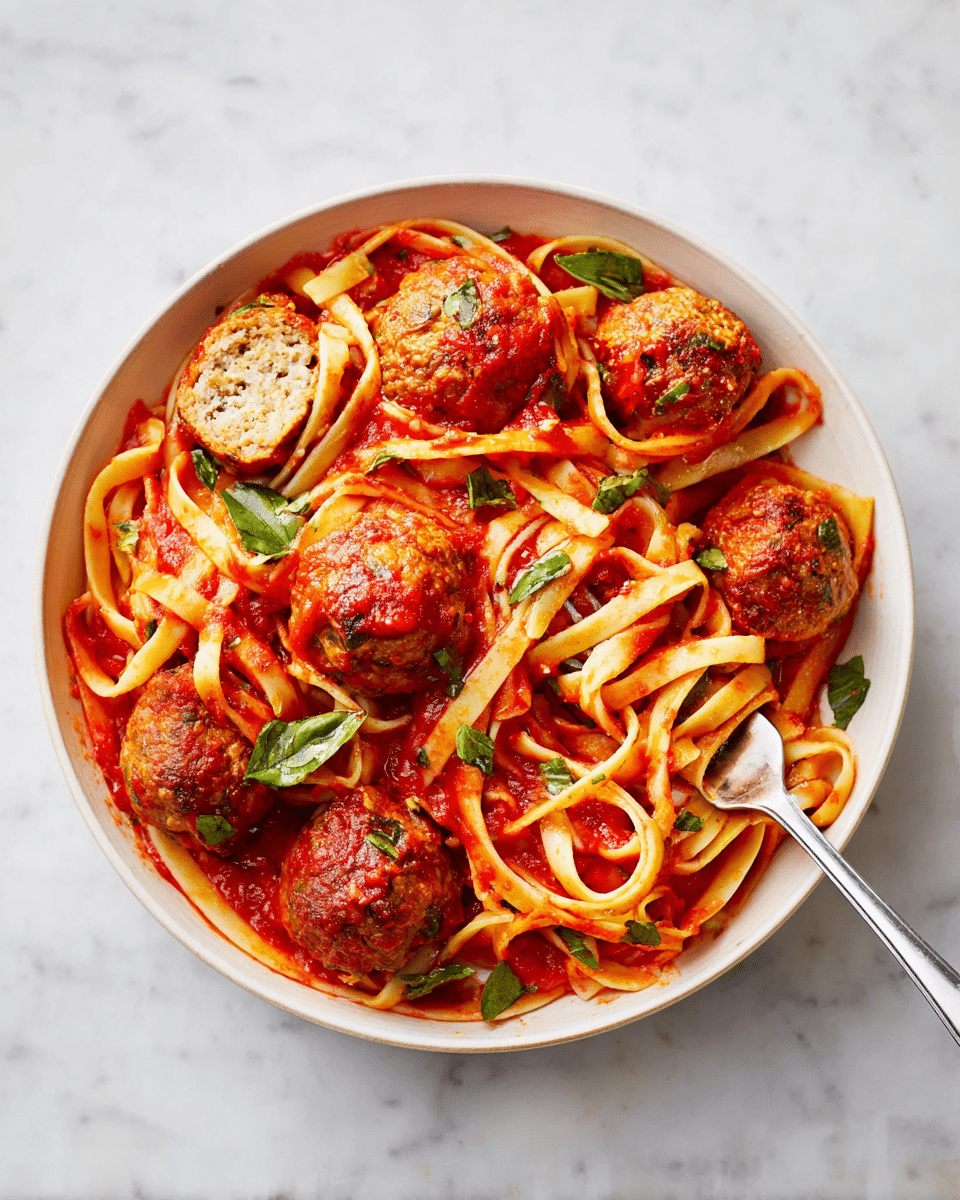 A white bowl filled with thick, flat linguine pasta coated evenly in bright red tomato sauce, mixed with six large, round meatballs that have a slightly crumbly texture. One meatball is cut open, revealing a light brown interior with herbs. Fresh green basil leaves are scattered on top, adding color contrast. A silver fork is twirling some pasta near the bottom right of the bowl. The bowl sits on a white marbled surface. photo taken with an iphone --ar 4:5 --v 7