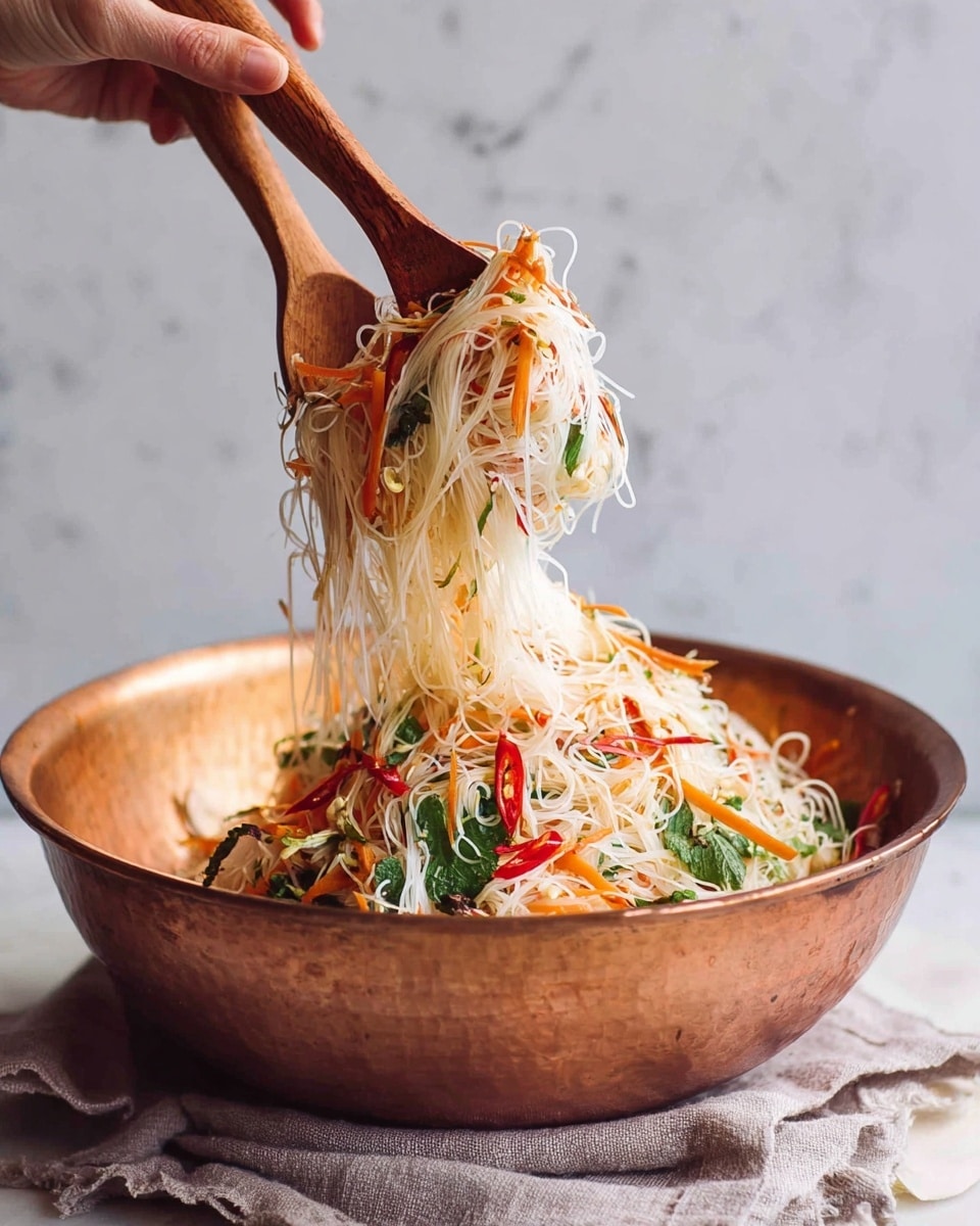 The image shows a large copper bowl filled with a colorful noodle salad. The salad has thin white rice noodles as the main layer, mixed with thin orange carrot strips, green herbs, and small slices of red chili peppers scattered throughout. Two wooden serving spoons are lifting the noodles above the bowl, showing the light and fresh texture of the salad. The bowl sits on a soft, light gray cloth on a white marbled surface. A woman's hand is holding one spoon, ready to serve the salad. photo taken with an iphone --ar 4:5 --v 7