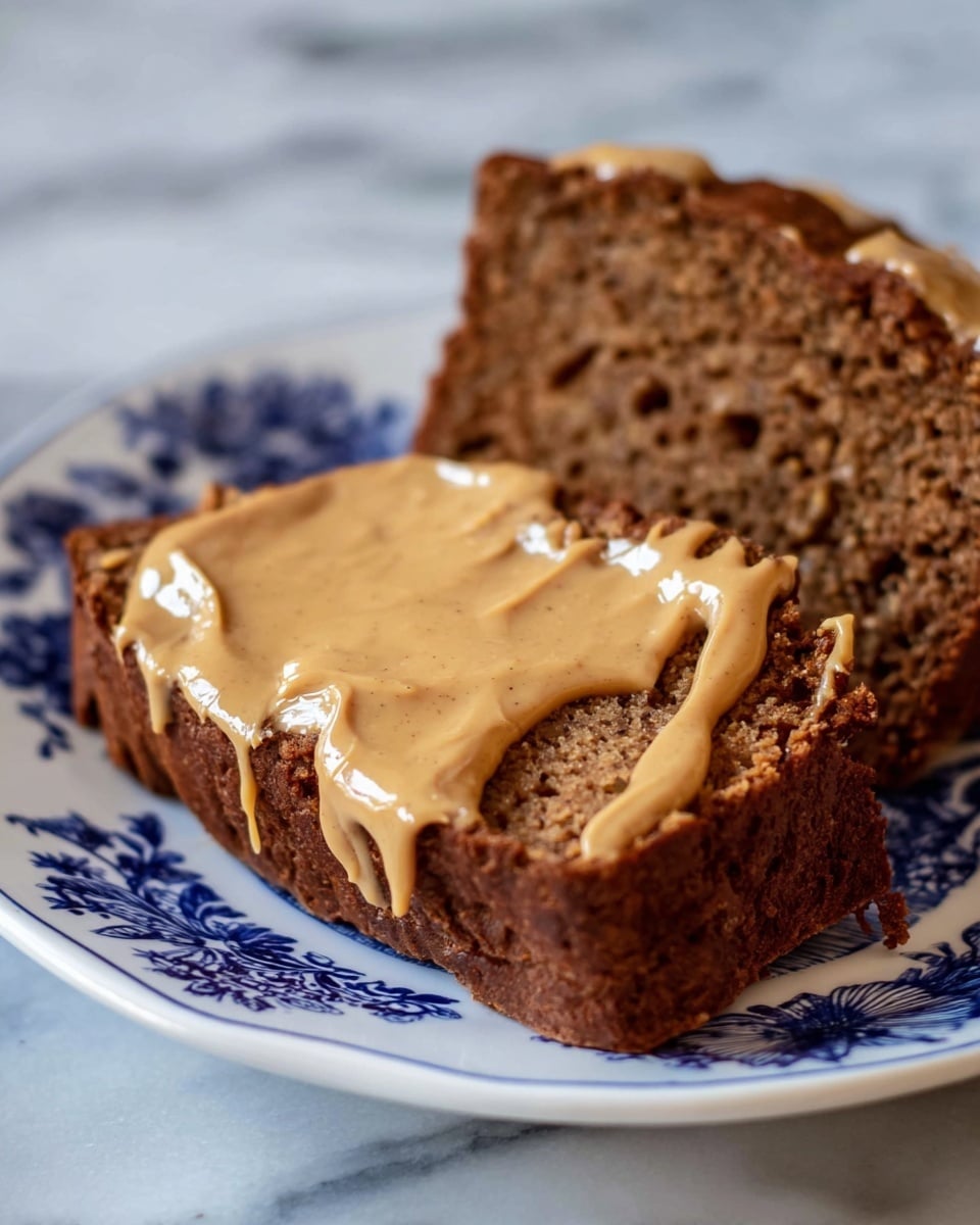 Two thick slices of dark brown banana bread rest on a white plate with blue floral patterns. The top slice is topped with a smooth, creamy layer of light brown peanut butter spread unevenly, creating a glossy texture with some drips on the side. The banana bread shows a dense, moist texture with small dark spots from the bananas. The scene is set on a white marbled surface. photo taken with an iphone --ar 4:5 --v 7