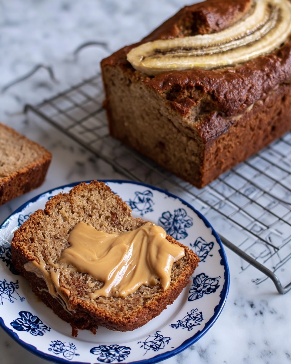 The image shows a loaf of banana bread on a cooling rack with a white marbled surface underneath, featuring a visibly moist, brown texture with two slices of banana on top. Next to the loaf, there is a white plate with a blue floral pattern holding two thick slices of the banana bread. The slice in front is covered with a light brown spread that looks creamy and smooth, drizzled unevenly across the top. The background is a white marbled texture. photo taken with an iphone --ar 4:5 --v 7