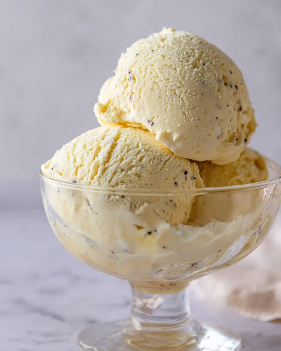 The image shows two large scoops of creamy vanilla ice cream placed one on top of the other in a clear glass dessert bowl. The ice cream is pale yellow with tiny black vanilla specks scattered throughout, giving it a textured look. The surface of the top scoop is smooth but slightly ridged from the scooping motion, while the bottom scoop looks softer and more uneven. The bowl is transparent with subtle reflections, sitting on a white marbled surface with a soft, plain background. Photo taken with an iphone --ar 4:5 --v 7
