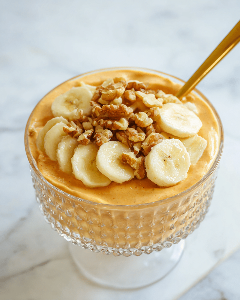 A clear glass bowl with a textured design holds a creamy, smooth orange base layer. On top, there is a layer of sliced banana pieces, pale yellow with a soft texture, scattered evenly. Mixed with the banana slices are small, uneven pieces of walnuts, light brown and adding a rough texture. A golden spoon is placed in the bowl, leaning slightly against the edge. The bowl is set on a white marbled surface. photo taken with an iphone --ar 4:5 --v 7