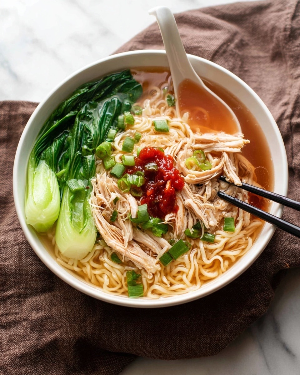 A white bowl filled with light brown broth at the base, topped with thin, light yellow noodles that fill most of the bowl. On one side, there is bright green bok choy lying flat over the noodles. In the center, shredded light brown chicken is placed over the noodles with a small dollop of red sauce and chopped green onions scattered on top. A white ceramic spoon rests inside the bowl on the left side, partially submerged in the broth, and a pair of black chopsticks hold some noodles on the right side. The bowl is placed on a soft, dark brown cloth over a white marbled surface. Photo taken with an iphone --ar 4:5 --v 7