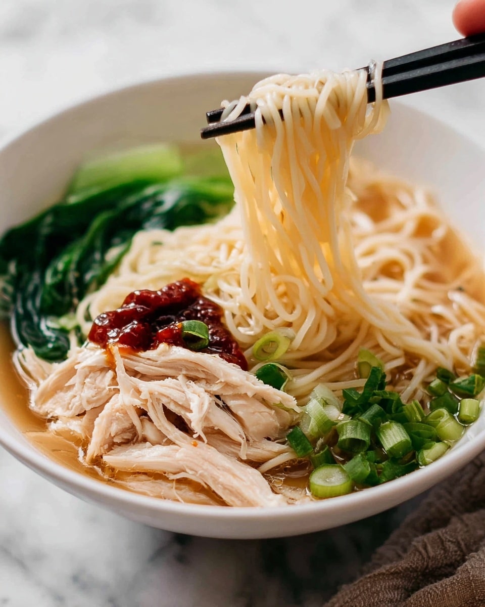 The image shows a bowl of noodle soup with four clear layers starting from the bottom: thin, pale yellow noodles soaking in light brown broth, soft white shredded chicken scattered over the noodles, bright green chopped spring onions and leafy greens placed on one side, and a dollop of dark red chili sauce on top of the chicken near the green onions. A pair of black chopsticks held by a woman's hand lifts a neat bundle of the noodles above the bowl. The bowl is white and rests on a white marbled surface. photo taken with an iphone --ar 4:5 --v 7