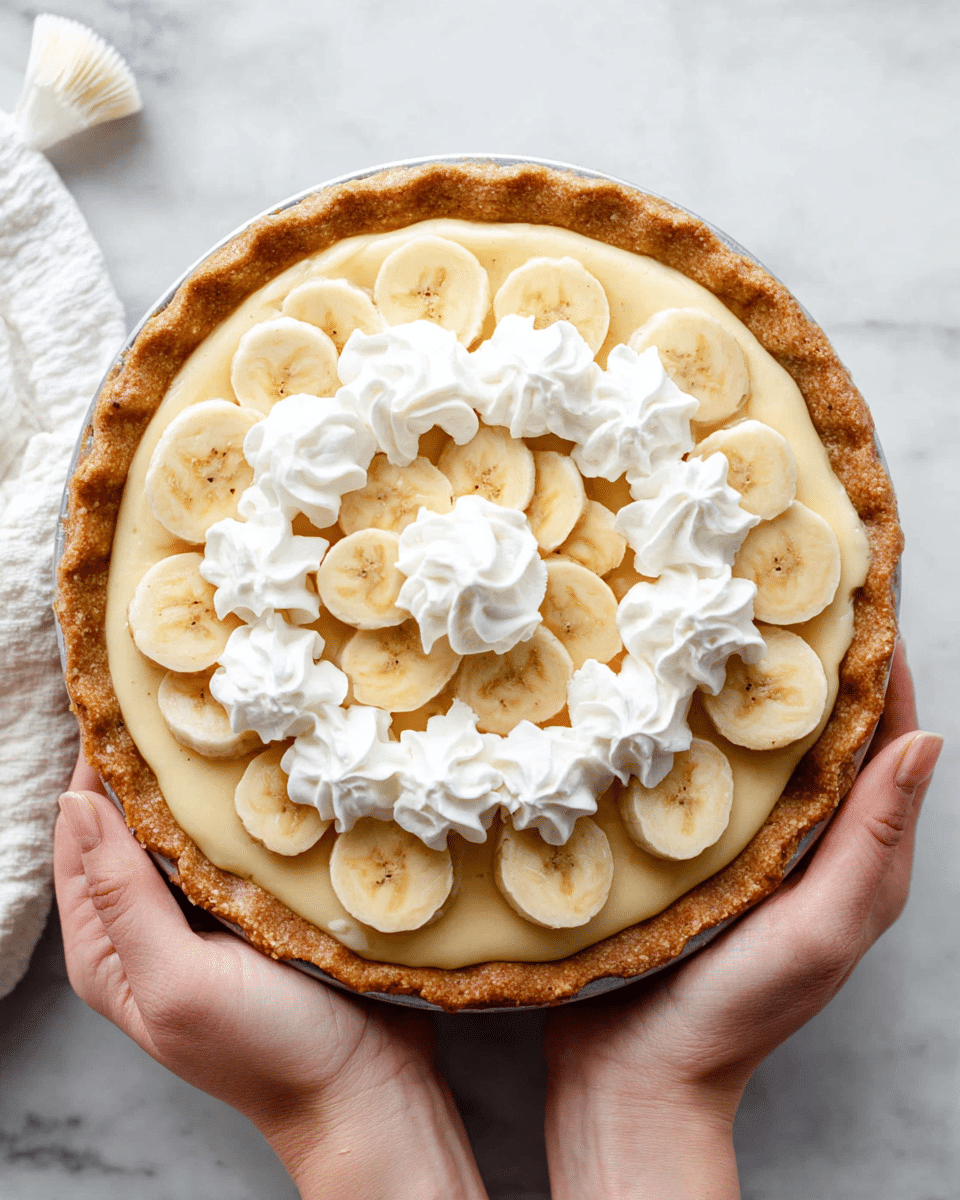 The image shows a banana cream pie held by two woman's hands, resting on a white marbled surface. The pie has a golden-brown crust that is thick and slightly crinkled around the edges. Inside, the pie features a smooth, pale yellow cream filling. On top of the cream, there is a circle of white whipped cream swirls, evenly spaced and thick. On top of the whipped cream layer, there are fresh banana slices arranged in two concentric circles, with each slice showing a light cream color with brown centers. A single banana slice sits on top of a small swirl of whipped cream in the center. A white piping bag is visible nearby on the white marbled surface. Photo taken with an iphone --ar 4:5 --v 7