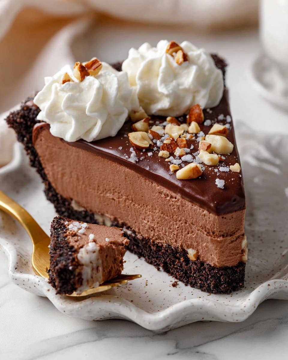 A slice of chocolate mousse pie with four visible layers sits on a white speckled plate with scalloped edges against a white marbled surface. The bottom layer is a dark, crumbly chocolate crust that forms the base and sides. Above it is a thick, creamy milk chocolate mousse layer with a smooth, airy texture. On top of the mousse sits a glossy, rich dark chocolate ganache layer, sprinkled with chopped hazelnuts and tiny flakes of salt. The pie slice is decorated with two swirls of white whipped cream placed near the crust edge. On the plate, a gold fork holds a bite-sized piece of the mousse layer with some crust. Photo taken with an iphone --ar 4:5 --v 7