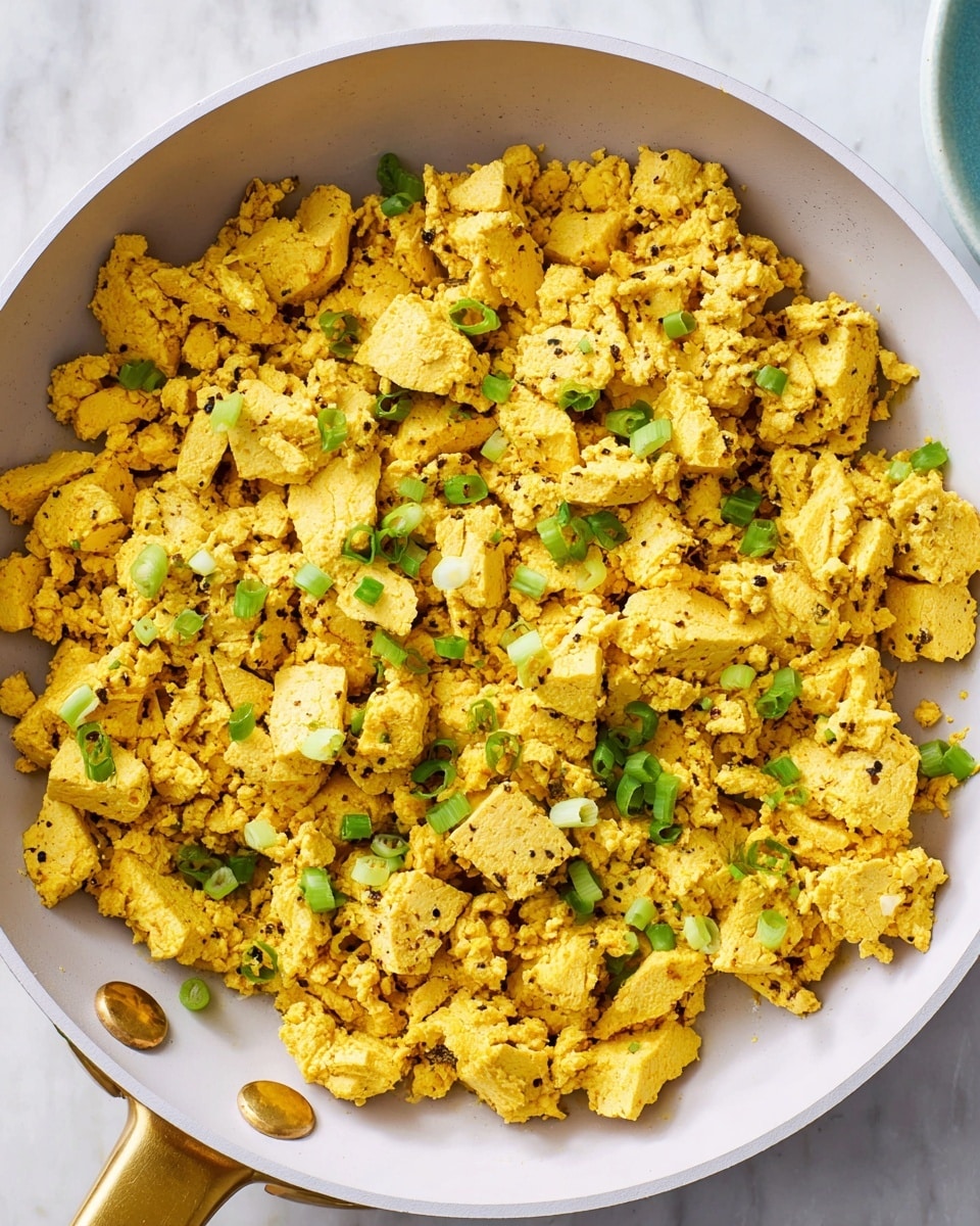 A close-up top view of a large white pan filled with scrambled pieces of scrambled tofu that are yellow and soft with a slightly rough texture. The tofu pieces are uneven in size and scattered around, sprinkled with small green rings of chopped green onion and tiny black pepper specks. The pan has two gold-colored rivets on the handle, and the whole scene is set against a white marbled surface. photo taken with an iphone --ar 4:5 --v 7