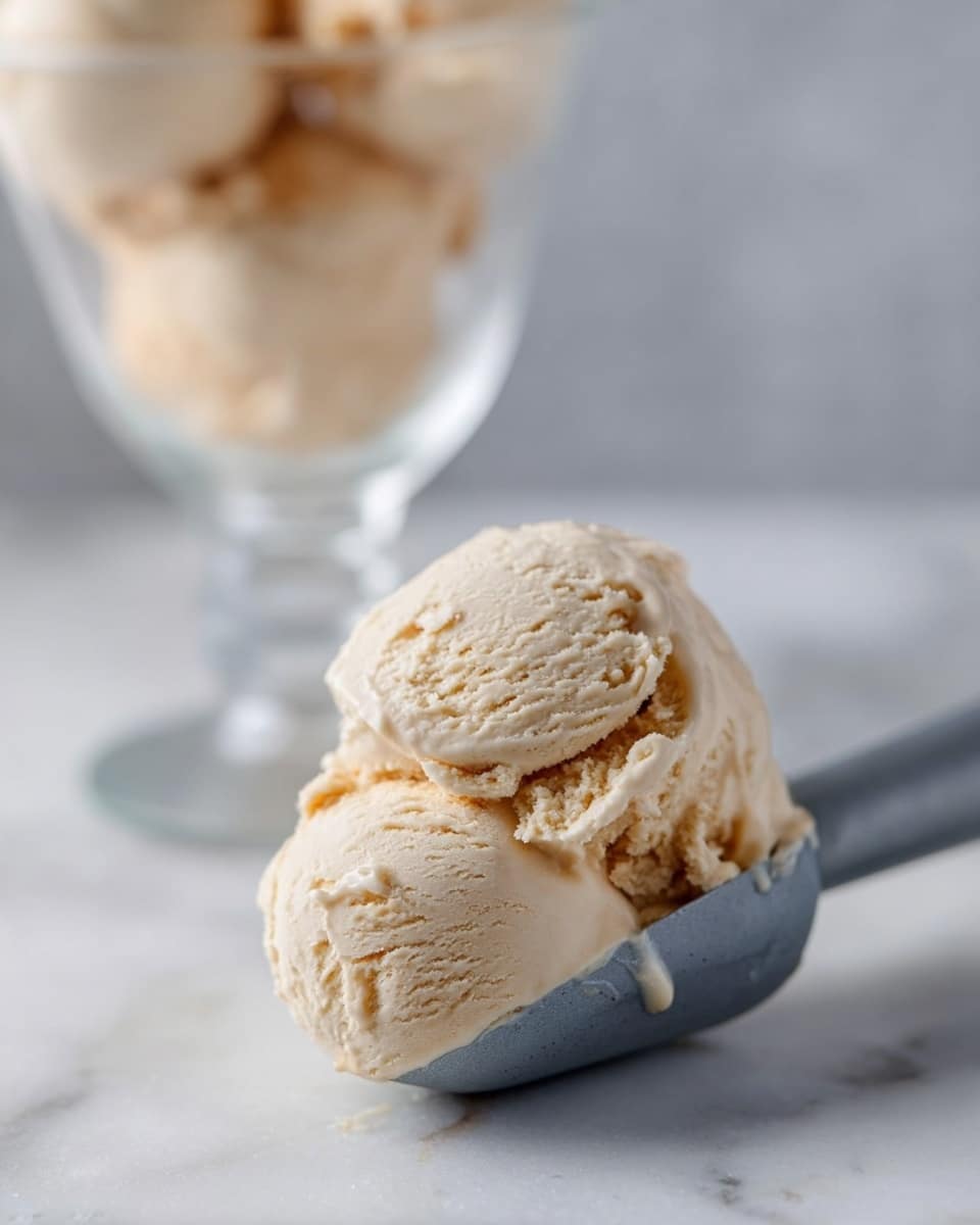 A clear glass dessert cup holds three large scoops of light beige ice cream with a smooth and creamy texture, slightly melting and dripping down the sides of the glass. The ice cream has subtle caramel swirls visible inside, adding contrast to the pale color. The cup sits on a white marbled surface, with a silver spoon placed nearby on the left side. The background is softly blurred and neutral-toned, highlighting the ice cream as the main focus. Photo taken with an iphone --ar 4:5 --v 7