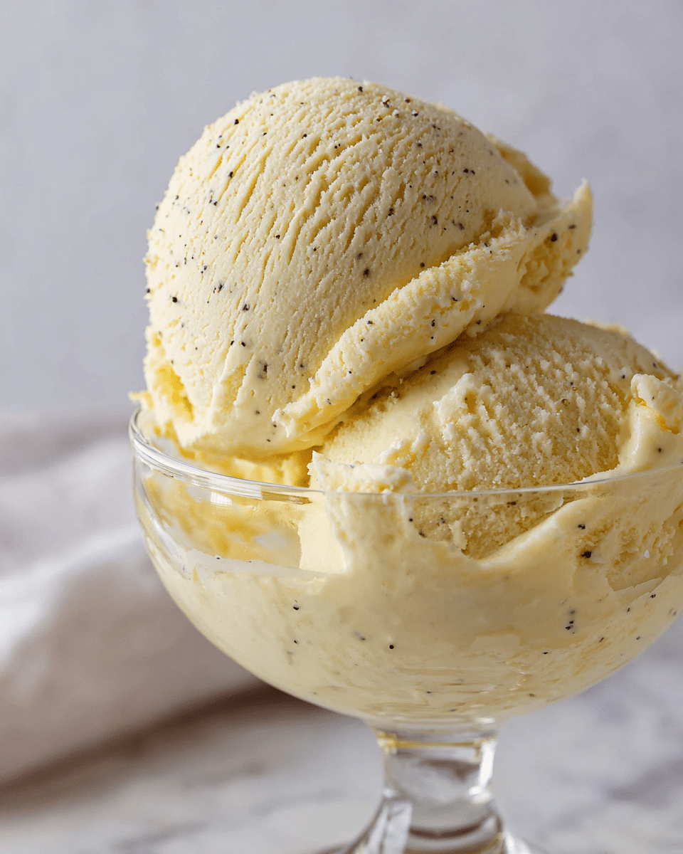 The image shows a clear glass dessert cup filled with two large scoops of creamy vanilla ice cream. The ice cream has a pale yellow color with tiny black vanilla bean specks scattered throughout. The texture is smooth and slightly melting around the edges, with some soft drips running down the inside of the glass. The background is blurred but appears to be a white marbled surface. photo taken with an iphone --ar 4:5 --v 7