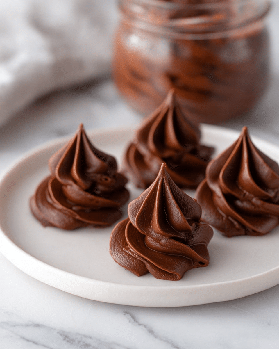 The image shows a white round plate holding five small swirls of rich, dark brown chocolate mousse, each with smooth, velvety texture and pointed peaks resembling rosettes. The mousse layers are piped thickly with a glossy finish, arranged with three in the front and two slightly blurred in the back. The plate sits on a white marbled surface, adding a soft, elegant background. A blurred jar filled with more chocolate mousse is visible behind the plate. photo taken with an iphone --ar 4:5 --v 7