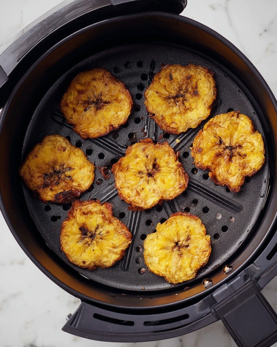 The image shows a black air fryer basket filled with eight golden-yellow fried plantain slices. Each plantain slice is round with a slightly rough and crispy texture, browned more in the center, and edges that are uneven and flaky. The basket has a perforated base to allow air flow, and it sits against a white marbled surface. The overall look is warm and crunchy, with the plantains spread loosely inside the basket. photo taken with an iphone --ar 4:5 --v 7