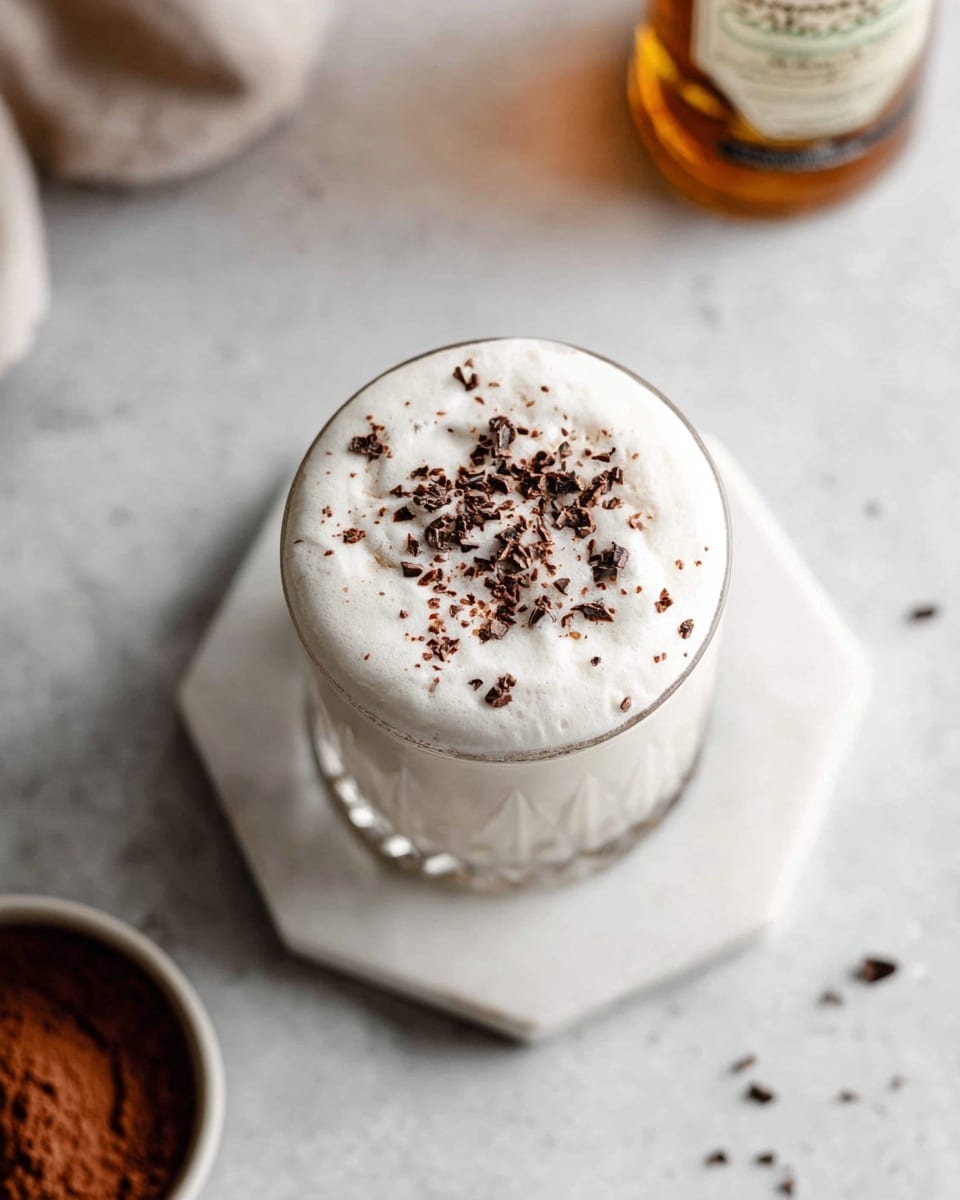 A close-up top view of a glass filled with a creamy, white frothy drink, layered with thick foam on top, sprinkled with small dark brown cocoa powder pieces scattered evenly over the foam. The glass sits on two stacked white marble hexagonal coasters, placed on a white marbled texture surface, with a blurred bottle and cocoa powder bowl in the background. Photo taken with an iphone --ar 4:5 --v 7
