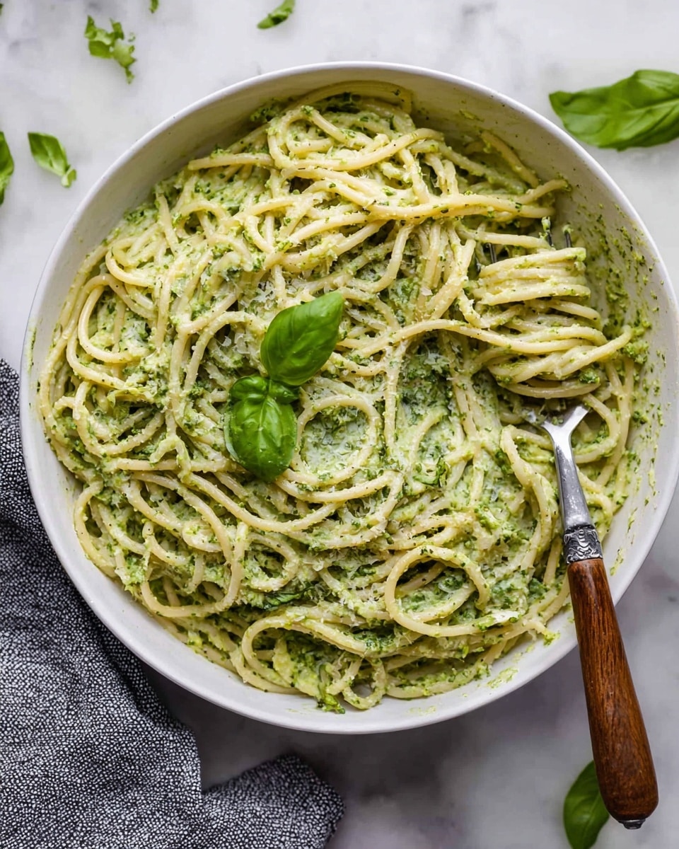 A large white bowl holds a serving of spaghetti coated in a creamy green pesto sauce, with the sauce having a slightly chunky texture from the herbs and nuts. The pale yellow pasta strands are mixed throughout the sauce, showing some loose curls and twists. A silver fork with a brown handle is lifting a twisted bunch of spaghetti near the right side of the bowl. A single fresh green basil leaf rests on top of the pasta in the center. The bowl sits on a white marbled surface with scattered basil leaves around it, and part of a gray textured cloth is visible on the left edge. Photo taken with an iphone --ar 4:5 --v 7