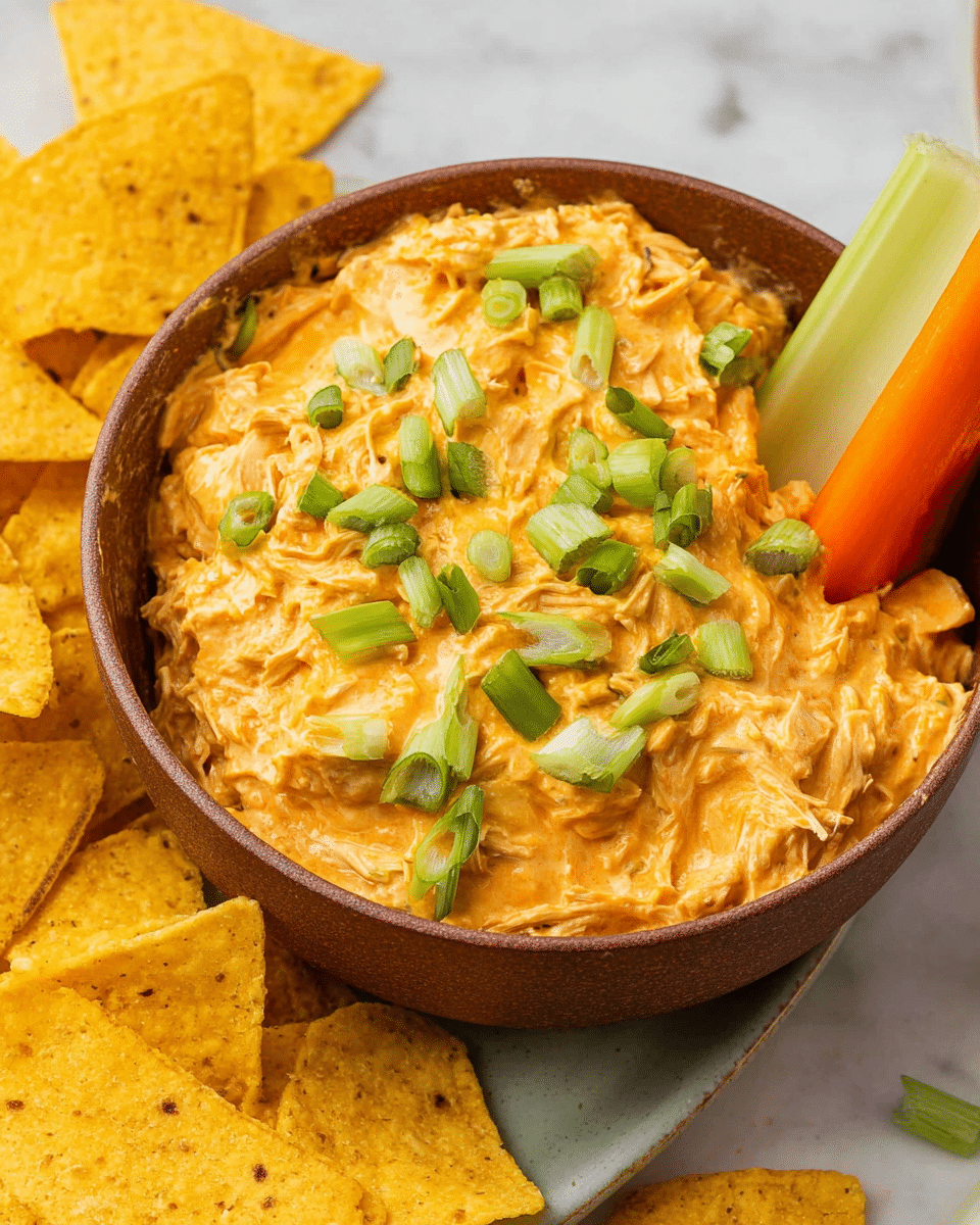 A brown bowl filled with a creamy, thick orange dip made from shredded chicken, topped with chopped green onions scattered on the surface. The dip looks soft and rich, covering the entire bowl with some chunks visible. A large white celery stick and orange carrot stick stand upright on the right side of the bowl, partially dipped. Several yellow tortilla chips surround the bowl, some dipped into the spread, showing texture and crispness. The background is a white marbled texture. photo taken with an iphone --ar 4:5 --v 7