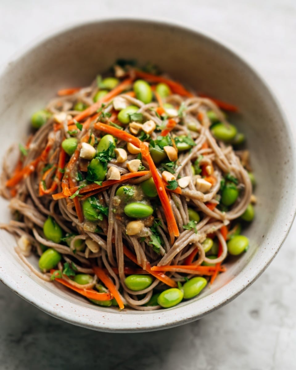 A close-up view of a white bowl filled with three main layers: the bottom layer has light brown soba noodles with a slightly rough texture; mixed within and on top there are bright green edamame beans and crisp, thinly sliced orange carrot strips; scattered throughout are small pieces of chopped peanuts and bits of fresh green herbs adding contrast. The bowl sits on a white marbled surface with a soft natural light highlighting the vibrant colors. photo taken with an iphone --ar 4:5 --v 7