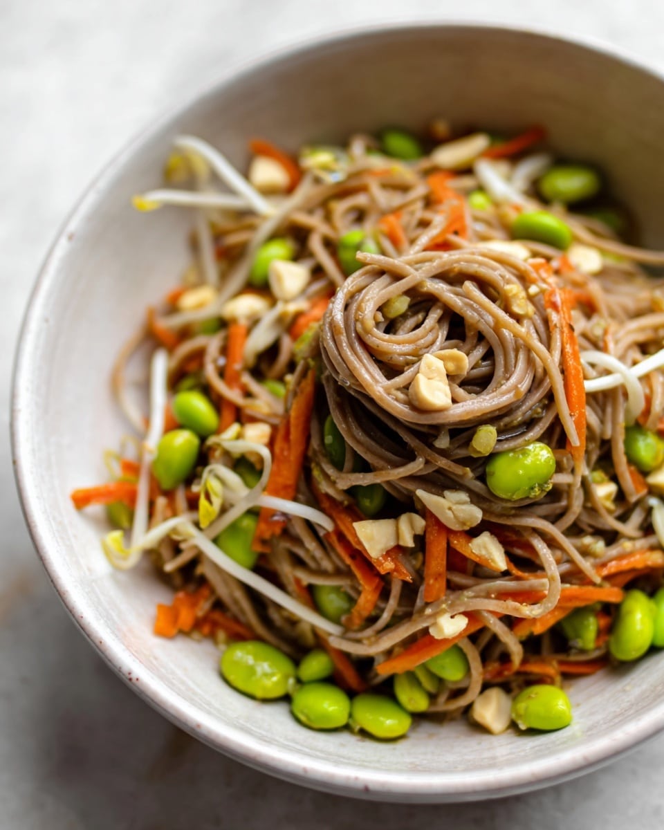 A close-up view of a white bowl filled with a noodle dish, featuring a thick bundle of light brown soba noodles twisted in the center. Around the noodles, there are bright green edamame beans, thin bright orange carrot strips, and white bean sprouts mixed evenly. Some small pieces of chopped nuts are scattered on top, adding texture. The bowl rests on a white marbled surface, and the photo highlights the freshness and mix of colors in the dish. photo taken with an iphone --ar 4:5 --v 7