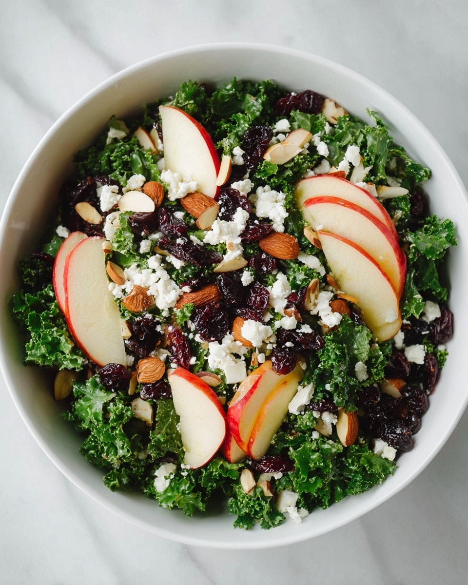 A white bowl filled with a fresh kale salad showing multiple layers: the bottom layer is dark green kale leaves with a curly texture, mixed with thin slices of red and white apple wedges placed evenly on top; scattered throughout are small, dark brown dried cherries adding a rich contrast, sprinkled with thin, light brown toasted almond slices, and dotted with small white crumbles of cheese, all set against a white marbled texture surface photo taken with an iphone --ar 4:5 --v 7