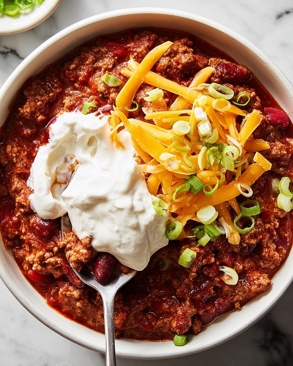 A close-up view of a bowl filled with chili, featuring a thick base layer of cooked ground meat mixed with kidney beans and rich red tomato sauce. On top, there is a layer of shredded bright orange cheddar cheese scattered unevenly. Small pieces of light-colored chopped onion and thin slices of green spring onions are sprinkled over the cheese. A spoon is scooping a part of the chili, lifting a dollop of smooth, creamy white sour cream that covers part of the cheese and chili. The bowl is white, placed on a surface with a white marbled texture. Photo taken with an iphone --ar 4:5 --v 7