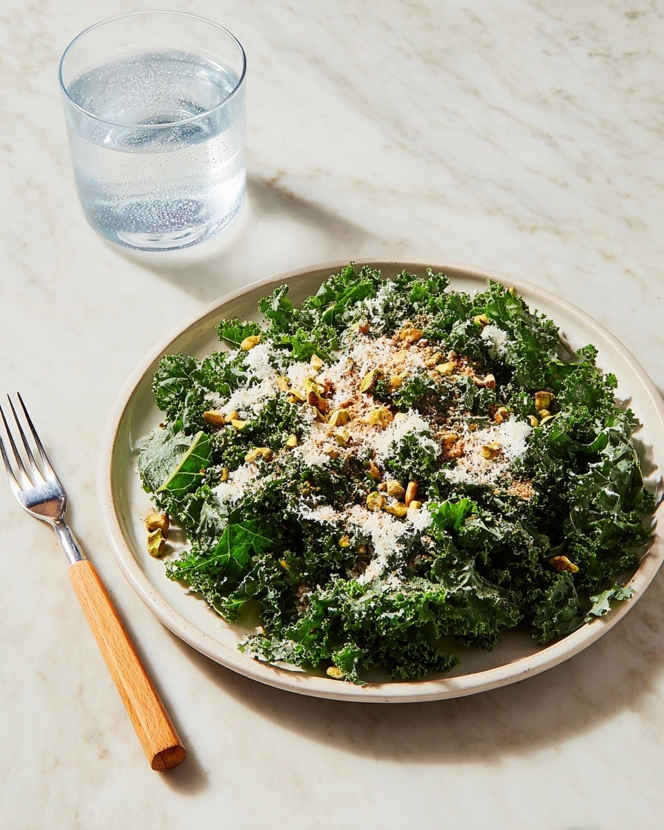 A white plate holds a fresh kale salad with curly, dark green leaves forming the base layer. On top, small pieces of chopped pistachios add a light green and brown crunchy texture, scattered evenly. A fine layer of grated white cheese is sprinkled over the salad, along with a light dusting of a brownish spice powder, giving contrast and texture variety. The plate sits on a white marbled surface. To the left of the plate, there is a fork with a light wooden handle and metal prongs, and above that, a clear glass filled halfway with sparkling water sits casting a subtle shadow. The photo taken with an iphone --ar 4:5 --v 7