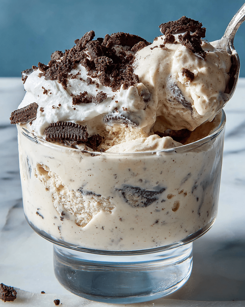 A clear glass dessert cup with a thick base holds a creamy, light beige ice cream layer mixed with small dark chocolate bits, topped with a fluffy white whipped cream layer, both sprinkled generously with dark chocolate cookie pieces. A silver spoon scoops up a mix of the base and whipped cream layers from the right side, showing the smooth and rich textures of the dessert. The scene is set on a white marbled surface with a soft blue background. photo taken with an iphone --ar 4:5 --v 7