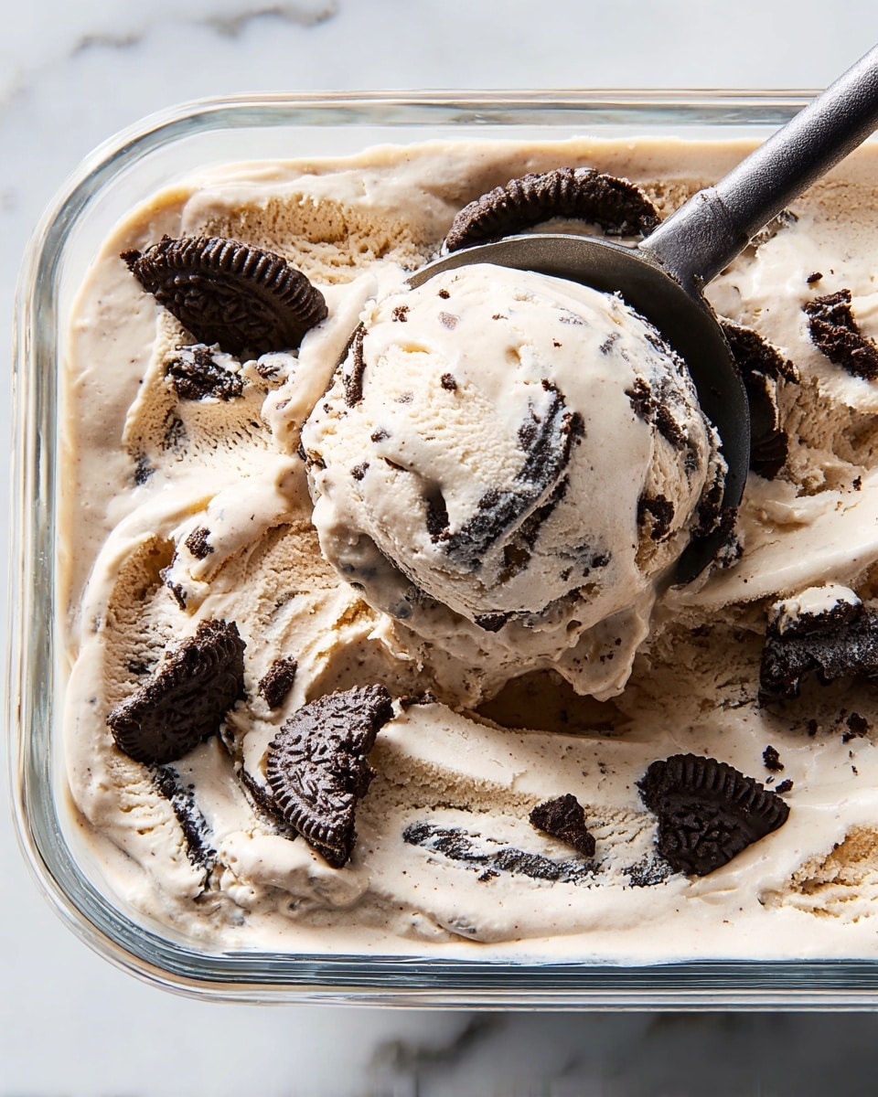 A close-up view of a single-layered creamy cookies and cream ice cream with a pale beige color mixed with dark brown chunks of cookie pieces, showing a thick, smooth, and soft texture inside a clear glass container; a dark metal spoon scoops up a generous portion of the ice cream, lifting it with visible cookie fragments embedded throughout, and scattered cookie pieces are resting on the ice cream surface; the scene is set against a white marbled texture. photo taken with an iphone --ar 4:5 --v 7