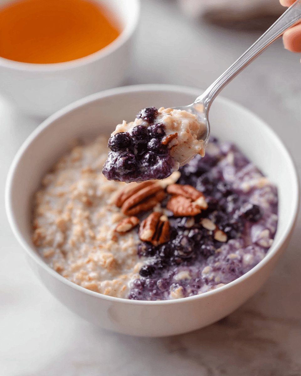 A white bowl sits on a white marbled surface, filled with a creamy oatmeal mixture that has two distinct layers: one side is a light beige with a smooth, dense texture, and the other side is a soft purple color with visible dark berries and crunchy pecan pieces scattered on top. A silver spoon held by a woman's hand lifts a scoop showing both the beige oatmeal and the purple oatmeal with pecans clearly visible. In the background, there is a blurred white bowl filled with an amber-colored liquid. Photo taken with an iphone --ar 4:5 --v 7
