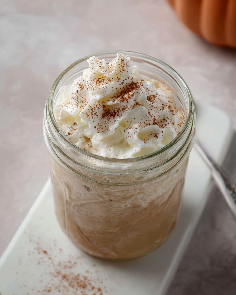 An open glass jar filled with a light brown creamy drink, topped with a generous swirl of white whipped cream that has a soft and fluffy texture, sprinkled with a fine dusting of brown cinnamon powder on top. The jar is placed on a white board, all set against a white marbled background. photo taken with an iphone --ar 4:5 --v 7