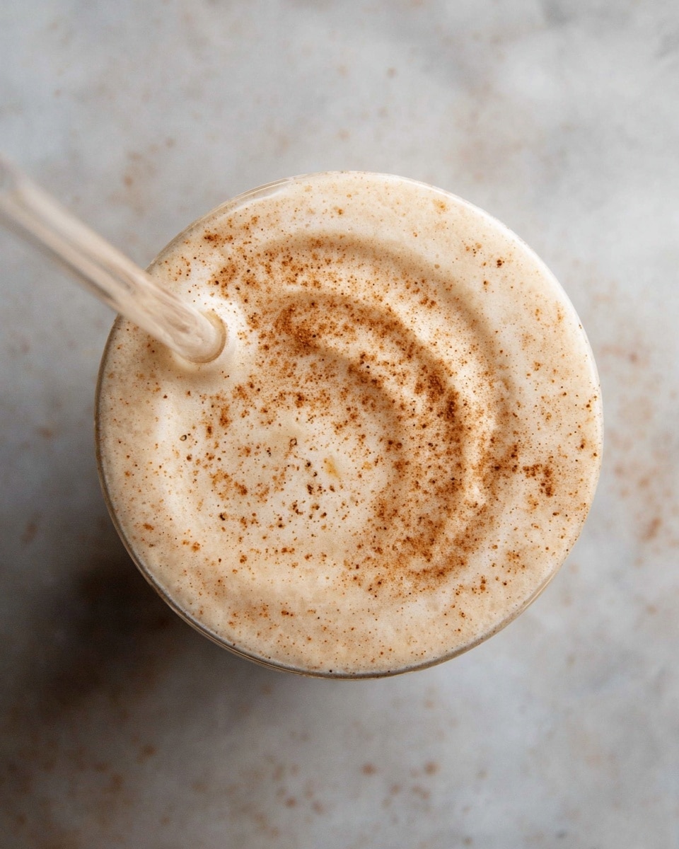 A close-up of a creamy beige drink in a clear glass seen from above, topped with a thin layer of froth swirled with light brown cinnamon powder, creating a smooth textured surface with small specks. A clear straw dips into the drink from the left side. The glass sits on a white marbled texture. photo taken with an iphone --ar 4:5 --v 7