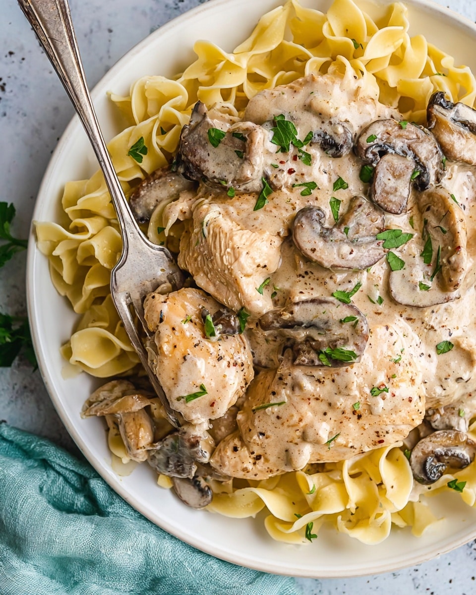 The dish shows a close-up of a white plate filled with wide egg noodles at the bottom, their pale yellow color visible and slightly glossy from sauce. On top, there are several pieces of creamy chicken coated in a thick light beige sauce with visible slices of dark brown mushrooms mixed in. The sauce appears smooth with specks of black pepper and small green parsley leaves scattered on top for garnish. A rustic silver fork is inserted in a scoop of noodles and chicken near the plate edge. The plate rests on a white marbled textured surface with a light teal cloth partially visible to the side. photo taken with an iphone --ar 4:5 --v 7