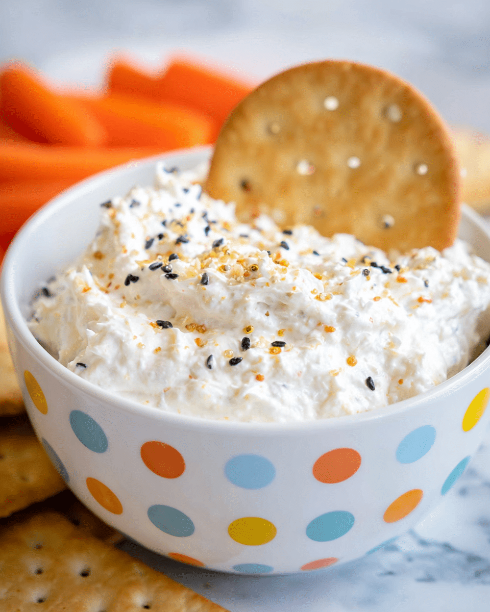 A close-up of a white bowl with colorful dots around the outside, filled with a thick, creamy white mixture that has a lumpy texture. The top is sprinkled with small black and golden-brown bits. A round, light brown cracker with small holes is partially dipped into the mixture from the back side. In the blurry background, small bright orange baby carrots can be seen. The bowl is placed on a surface with a white marbled texture photo taken with an iphone --ar 4:5 --v 7