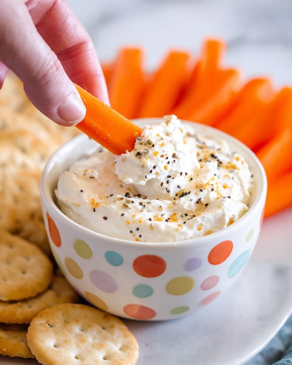 A close-up of a white bowl with colorful polka dots filled with creamy white dip mixed with small black and brown seasoning flakes. A bright orange carrot stick is dipped into the thick dip, held by a woman's hand with visible fingernails. Behind the bowl, more carrot sticks and round light tan crackers are placed, all resting on a white marbled surface. The image focus is sharp on the dip and carrot stick, showing texture and seasoning clearly. photo taken with an iphone --ar 4:5 --v 7