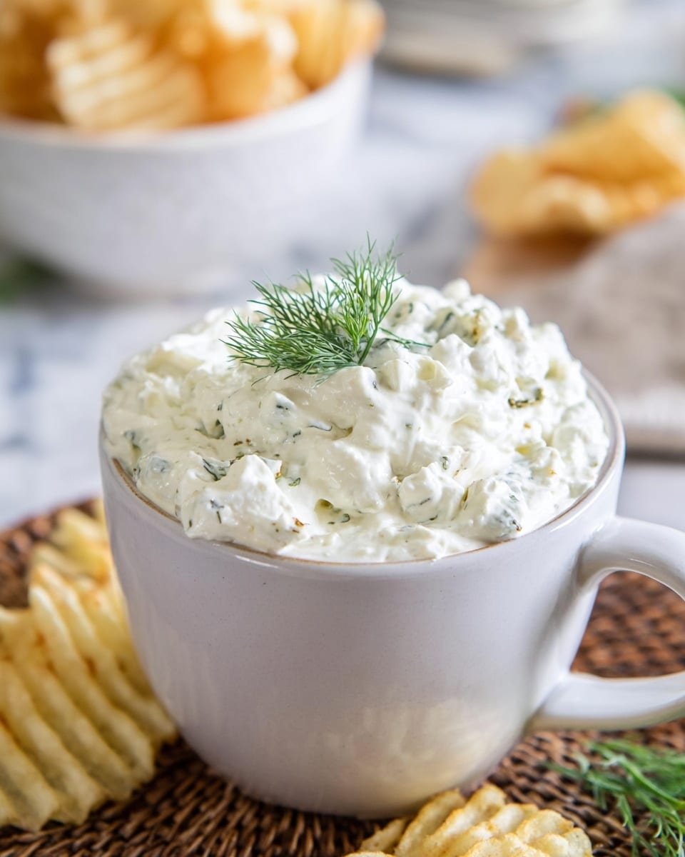 A white mug filled with a thick, creamy white mixture with small curds, lightly speckled with green herbs, is topped with a small fresh green dill sprig. The mug sits on a woven mat on a white marbled surface. In the background, out of focus, is a white bowl filled with crinkle-cut potato chips and some more chips placed on the white marbled surface near the mug's base. The photo has soft, natural lighting and a close-up view of the creamy dip. photo taken with an iphone --ar 4:5 --v 7
