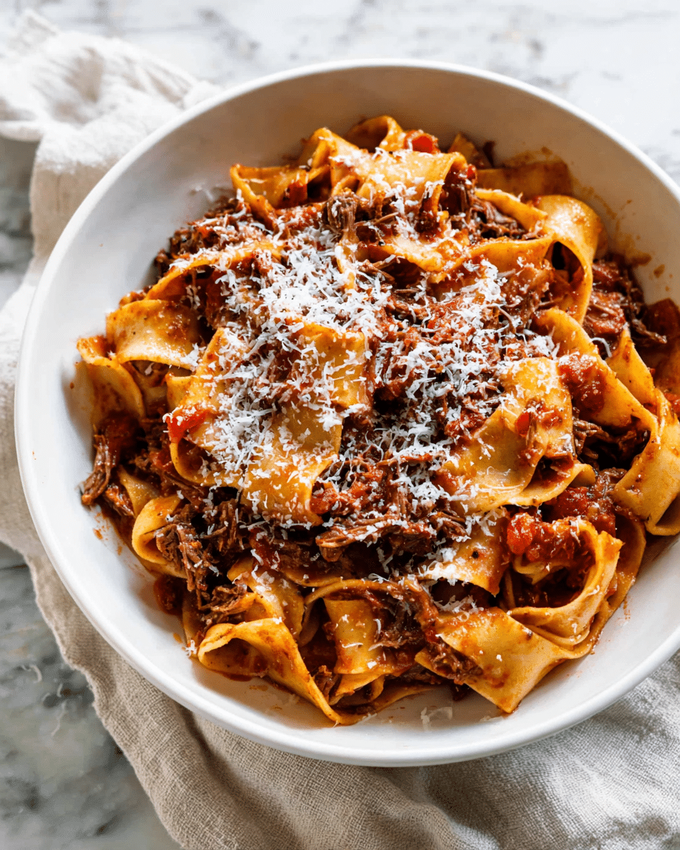 A white bowl filled with wide, flat pasta noodles coated in a rich, reddish-brown meat sauce with visible chunks of shredded beef and diced tomatoes. The pasta is layered thickly, with the sauce sitting evenly among the noodles, giving a glossy texture. On top, there is a generous sprinkle of finely grated white cheese, adding a light contrast to the darker sauce and noodles. The bowl sits on a white marbled surface with a soft cloth partially visible underneath it. photo taken with an iphone --ar 4:5 --v 7