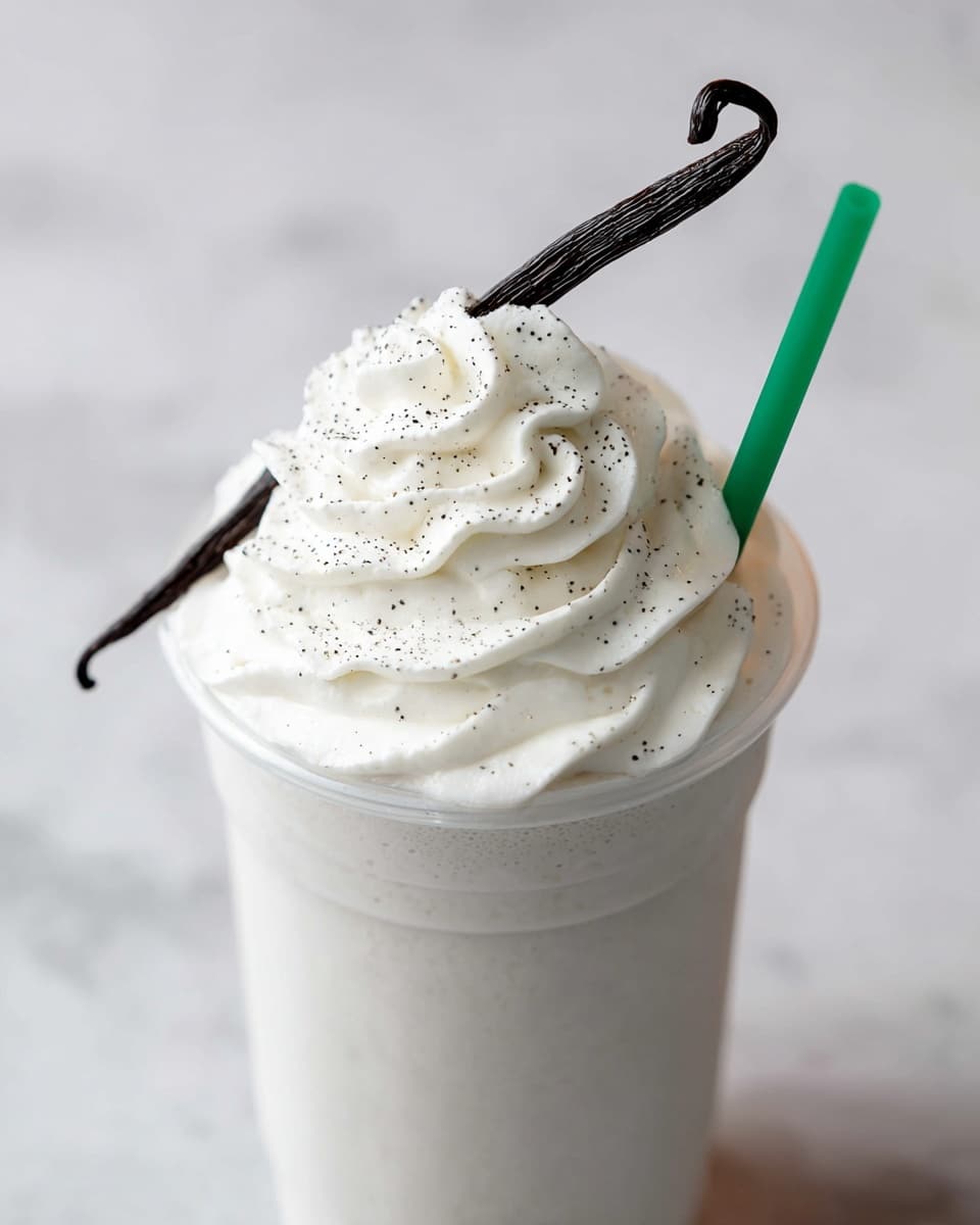A close-up view of a creamy vanilla milkshake in a clear cup, topped with a thick layer of white whipped cream sprinkled with tiny black vanilla bean specks, and a single whole dark brown curved vanilla bean placed vertically on top. A green straw stands on the right side, slightly sunk into the whipped cream. The cup is set on a white marbled surface that adds a soft background texture. photo taken with an iphone --ar 4:5 --v 7