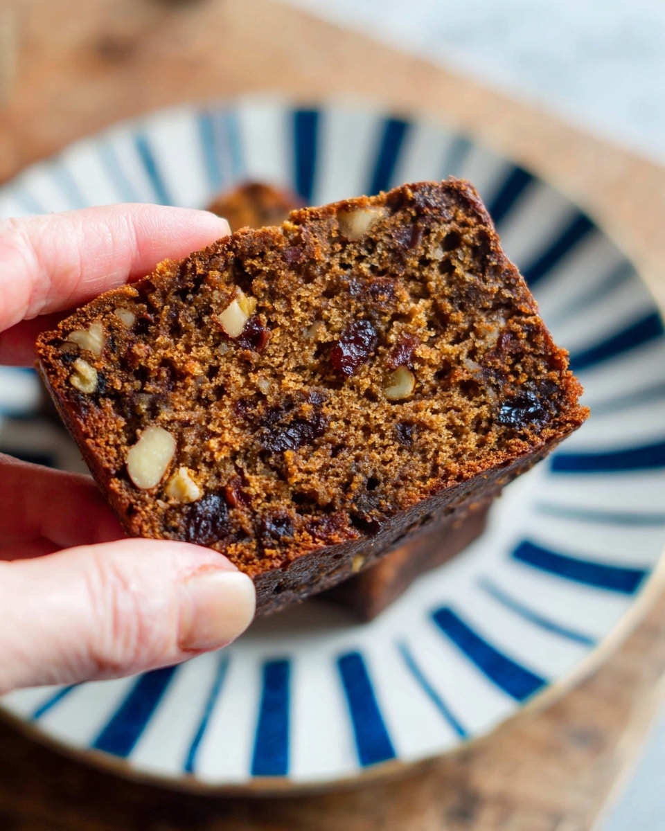 A close-up image of a single slice of dark brown fruit and nut loaf, held by a woman's hand on the left side of the frame. The loaf slice shows a dense texture with visible pieces of nuts and dark dried fruits spread unevenly inside. The slice has a slightly rough top crust and appears moist. The background shows parts of a white plate with blue stripes underneath the slice, resting on a wooden surface, now described as a white marbled texture. Photo taken with an iphone --ar 4:5 --v 7