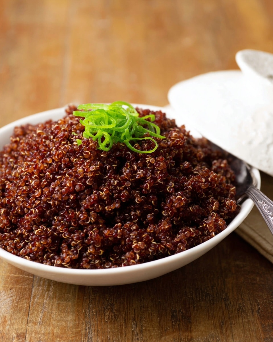 A large mound of cooked red quinoa with a glossy, slightly textured look fills a white bowl, topped with a small pile of chopped green onions that add a bright, fresh contrast in color and texture. The bowl is placed on a wooden surface, and a white textured lid leans against the bowl on the right side. A silver spoon with a simple handle is partially visible, resting inside the bowl behind the quinoa. Photo taken with an iphone --ar 4:5 --v 7