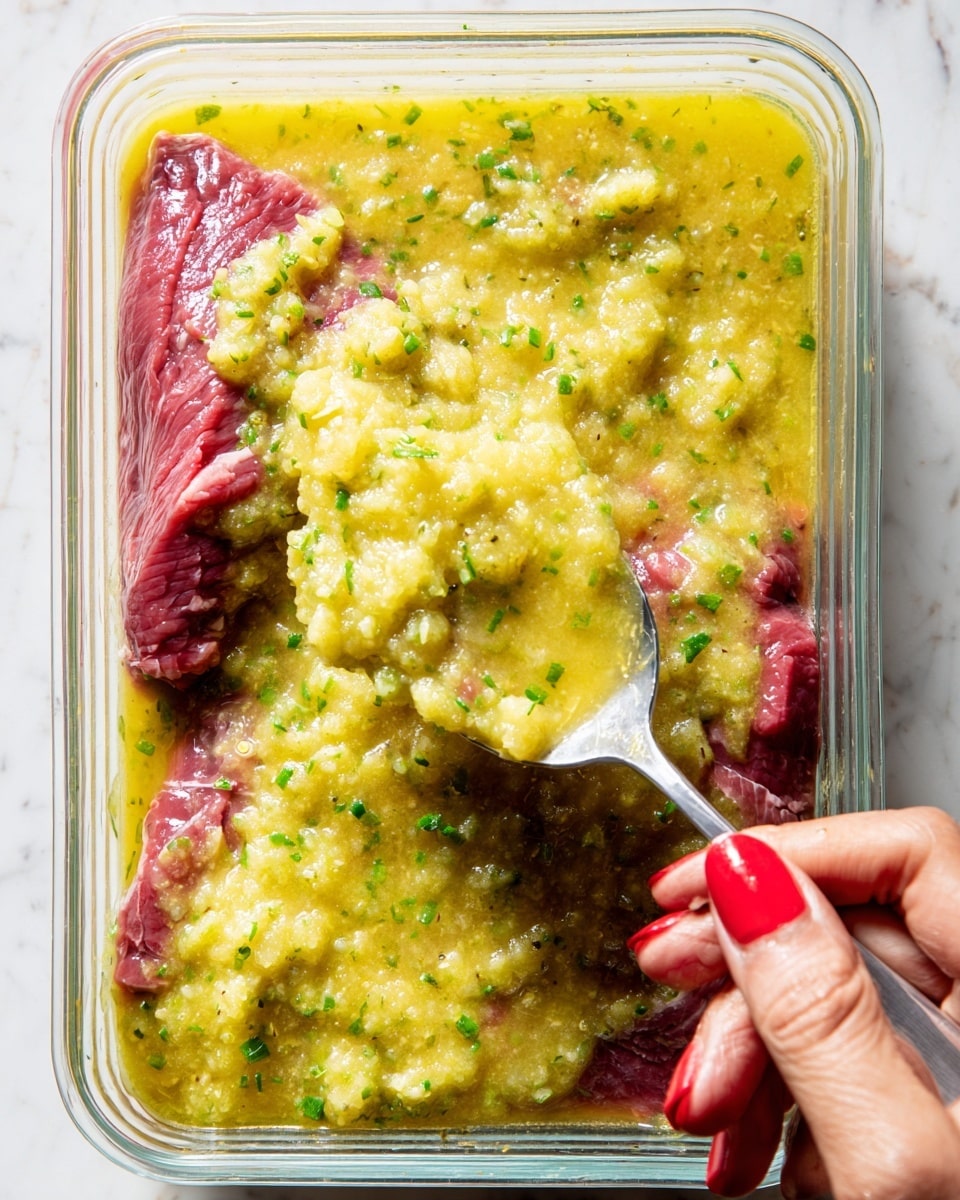 A close-up view of a layered dish in a clear rectangular glass container, showing two layers: the bottom layer is red raw beef, with visible texture and marbling, and the top layer is a chunky yellow marinade with small green herb bits spread unevenly over the meat. A woman's hand with red nail polish holds a silver spoon, scooping and spreading the thick, coarse mixture on top of the beef. The whole scene is set against a white marbled texture background. photo taken with an iphone --ar 4:5 --v 7