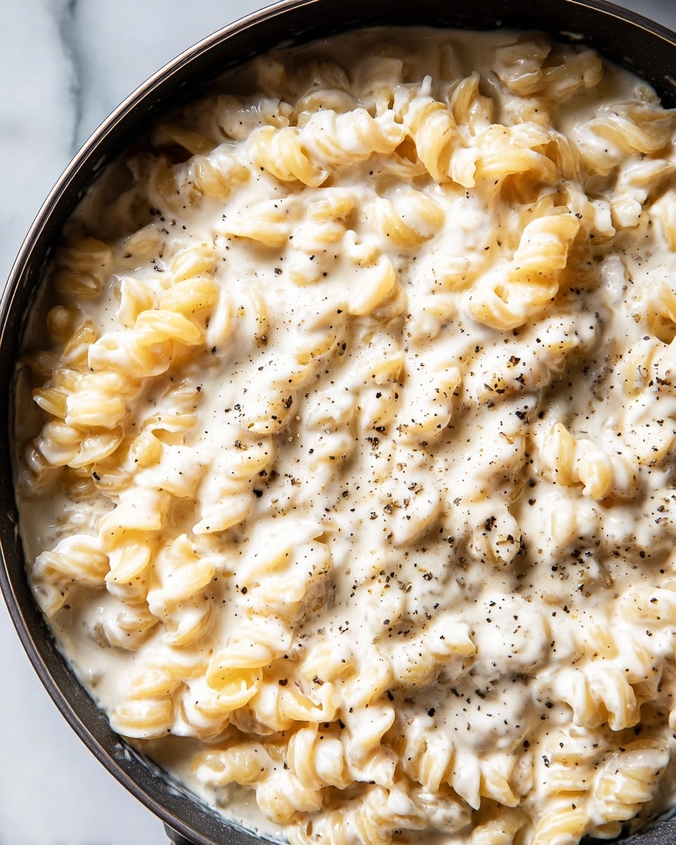 A close-up view of a large black pan filled with creamy white sauce-covered pasta. The pasta is short and curly, evenly coated with a thick, smooth white sauce sprinkled with small black pepper bits. The pasta pieces are tightly packed, and the sauce has a glossy texture, pooling slightly around the edges. The background surface is a white marbled texture. photo taken with an iphone --ar 4:5 --v 7