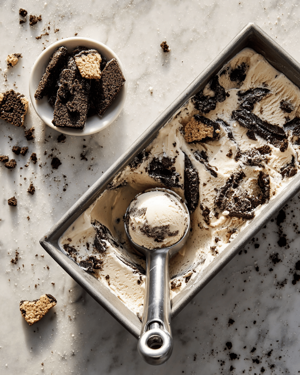 The image shows a metal container filled with creamy white ice cream mixed with chunks and crumbs of dark brown chocolate cookies, creating a layered and textured look with specks and larger cookie pieces spread unevenly on top. A large metal ice cream scoop holds a round ball of this cookies and cream ice cream near the center. To the left, a small white bowl holds more broken cookie pieces. The scene is set on a white marbled surface, and there are cookie crumbs scattered around the container and bowl. Photo taken with an iphone --ar 4:5 --v 7