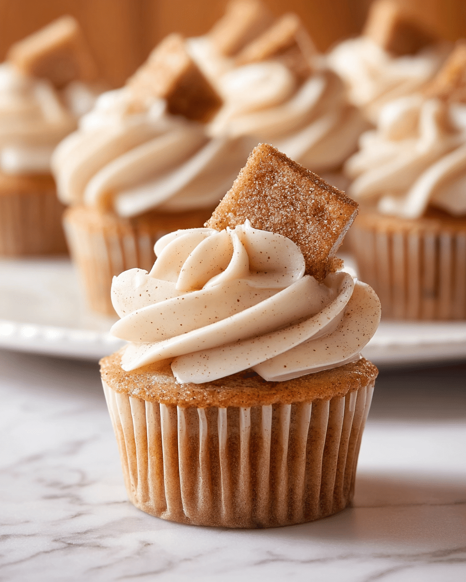 A close-up of a single cupcake with a light brown base wrapped in a ridged paper liner, topped with smooth, creamy swirls of pale beige frosting with tiny specks, and crowned by a small square piece of cinnamon sugar-coated cake placed at an angle on top. Behind it, more cupcakes with similar frosting are partially visible on a white plate, all resting on a white marbled surface. photo taken with an iphone --ar 4:5 --v 7