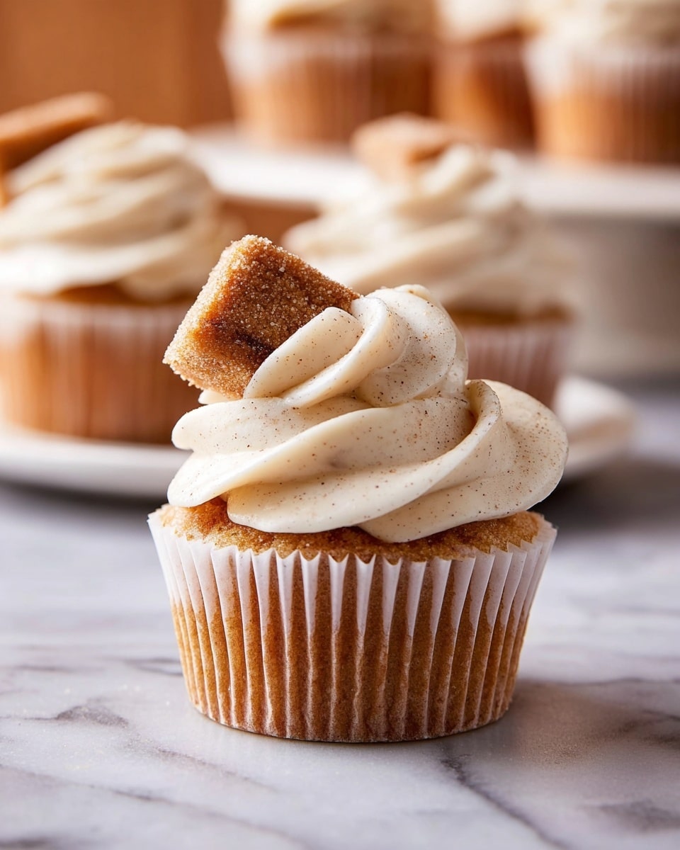 A cupcake with a light brown base wrapped in a white paper liner sits on a white marbled surface. On top is a generous swirl of creamy, off-white frosting with tiny specks, giving it a smooth and fluffy texture. Stuck at an angle in the frosting is a small rectangular piece of cinnamon sugar-coated cake, light brown with a slightly rough texture. In the background, more similarly frosted cupcakes rest on a white plate on the same white marbled surface. photo taken with an iphone --ar 4:5 --v 7