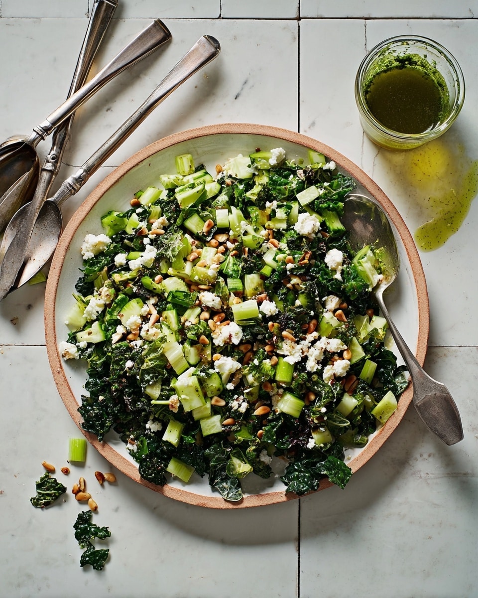 A large white plate holds a fresh mixed salad with three visible layers: the bottom layer is made of dark green kale leaves with a rough texture, the middle layer has light green chopped celery and cucumber pieces with a crisp texture, and the top layer is sprinkled with white crumbled cheese and small brown toasted pine nuts, all lightly drizzled with green pesto sauce. The plate is placed on a white marbled tile surface, next to three silver salad servers and a small glass cup filled with green pesto sauce. Some scattered bits of salad are around the plate, adding a natural, casual look. Photo taken with an iphone --ar 4:5 --v 7