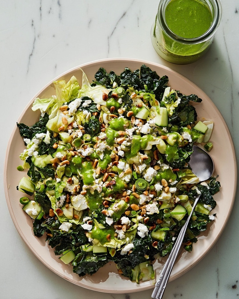 A large white plate holds a fresh salad with multiple layers: the bottom layer has dark green kale leaves mixed with light green chopped cabbage, followed by medium green diced cucumber cubes scattered on top, and sprinkled generously with small white crumbly cheese pieces. Light brown toasted pine nuts are spread evenly across the salad. Bright green sliced scallions add more color, and dollops of green herb dressing are drizzled over the top. A silver serving spoon rests on the right side of the plate. The plate sits on a white marbled surface next to a small glass jar filled with green dressing. photo taken with an iphone --ar 4:5 --v 7