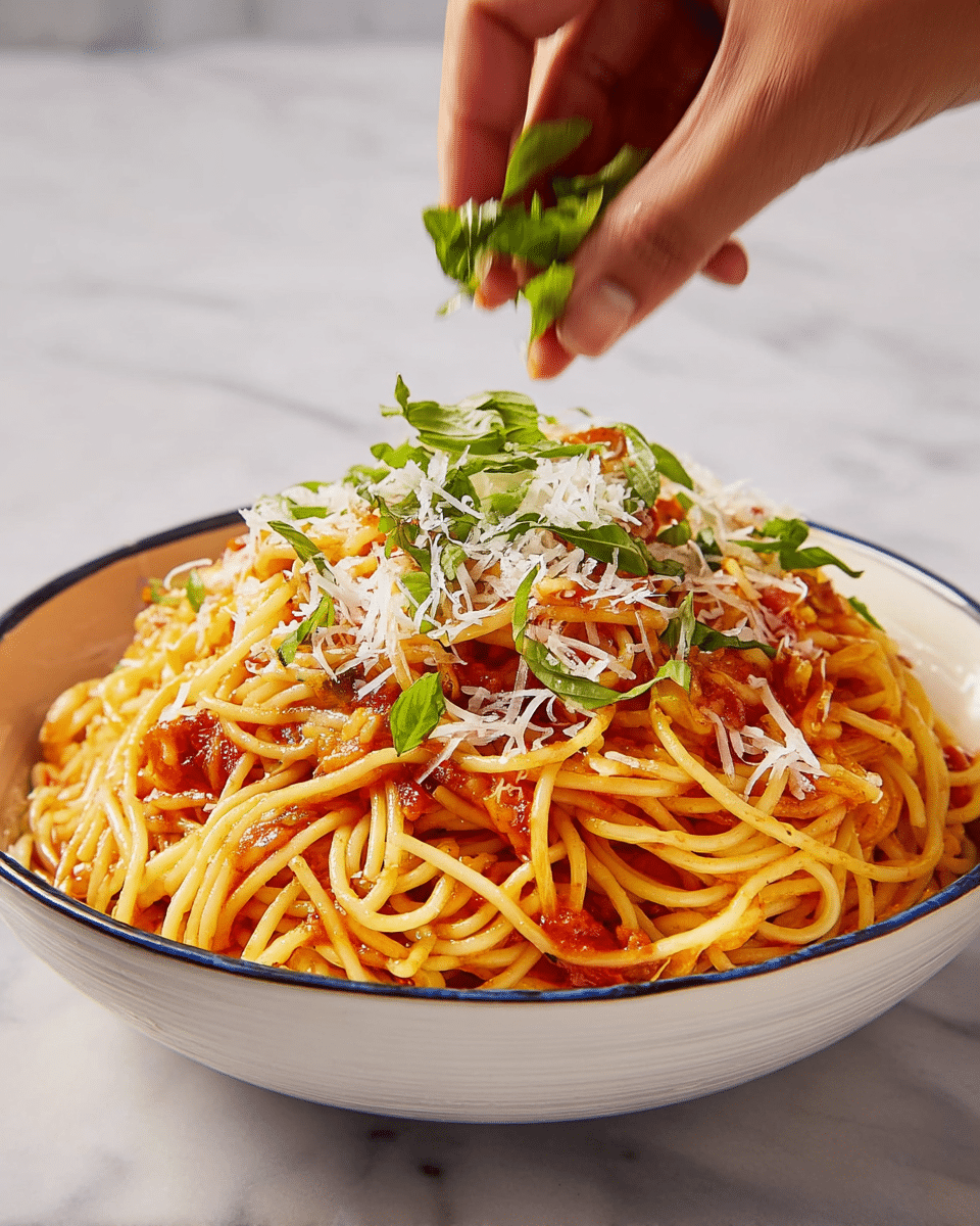 A large white bowl filled with a big pile of yellow spaghetti, mixed with red tomato sauce and small bits of tomato and onion scattered throughout. The top layer of spaghetti is sprinkled with shredded white cheese and thin strips of fresh green basil leaves. A woman's hand is above the bowl, adding more green basil. The bowl sits on a white marbled surface. photo taken with an iphone --ar 4:5 --v 7