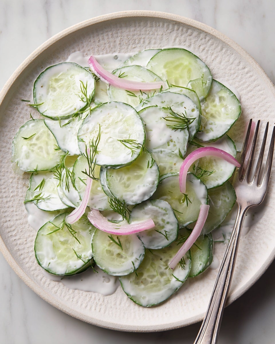 The image shows a salad made of thin cucumber slices mixed with a white creamy dressing, spread on a single layer over a white textured plate. There are a few thin pink onion slices scattered among the cucumbers, and small green dill sprigs are sprinkled on top, adding a fresh touch. A silver fork is positioned on the right edge of the plate, resting on the clean white marbled surface. Photo taken with an iphone --ar 4:5 --v 7