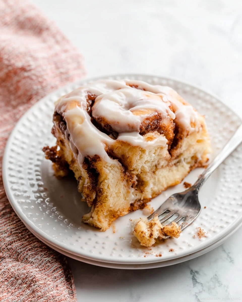 A slice of cinnamon roll with several thick, soft layers of light brown dough twisted with darker cinnamon filling sits on a white plate with a raised dot pattern near the rim. The roll is topped with a glossy, white glaze that drips slightly down the sides, adding a shiny texture. Small crumbs from the roll scatter on the plate, and a silver fork with traces of cinnamon sugar rests beside the slice. The scene is set on a white marbled surface with a pink cloth partially visible in the lower left corner. Photo taken with an iphone --ar 4:5 --v 7